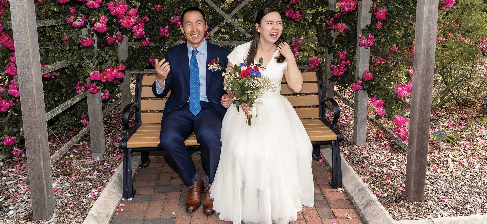 A bride and groom laugh while sitting on a bench surrounded by flowers