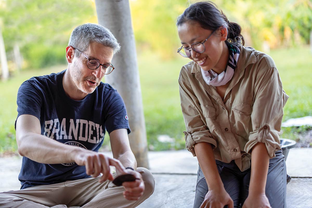 Charles Golden, wearing a Brandeis t-shirt, points to an object. Grace Horseman smiles at the object.