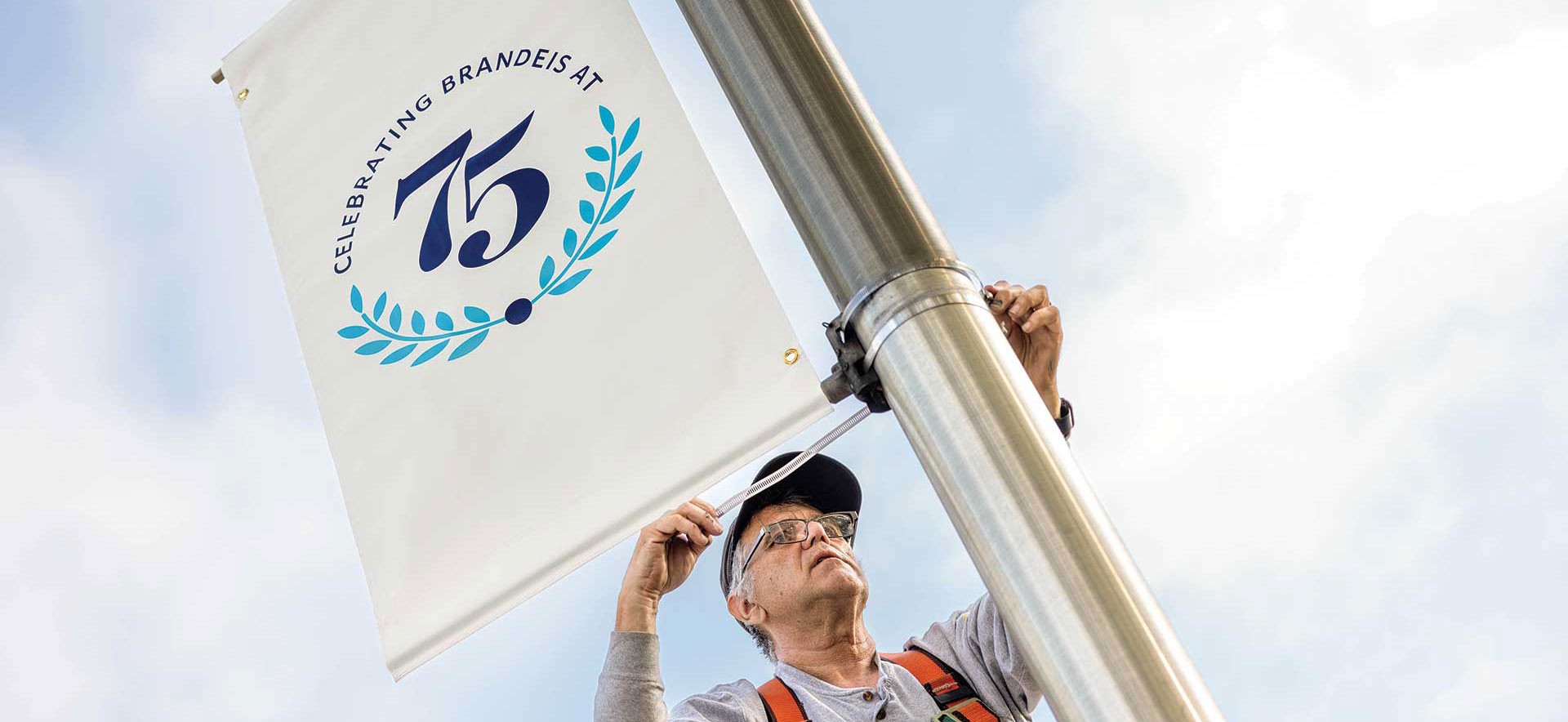 A facilities employee hangs a Brandeis 75th Anniversary Banner on a light post.