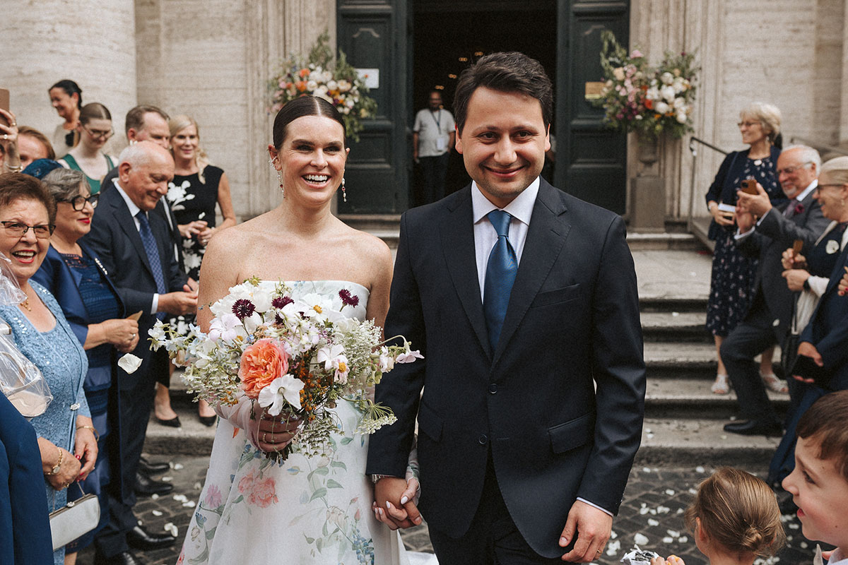 A bride and groom hold hands as wedding guests applaud.