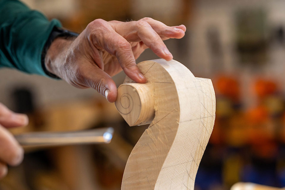 Hands carving a wooden cello's scroll with a chisel.