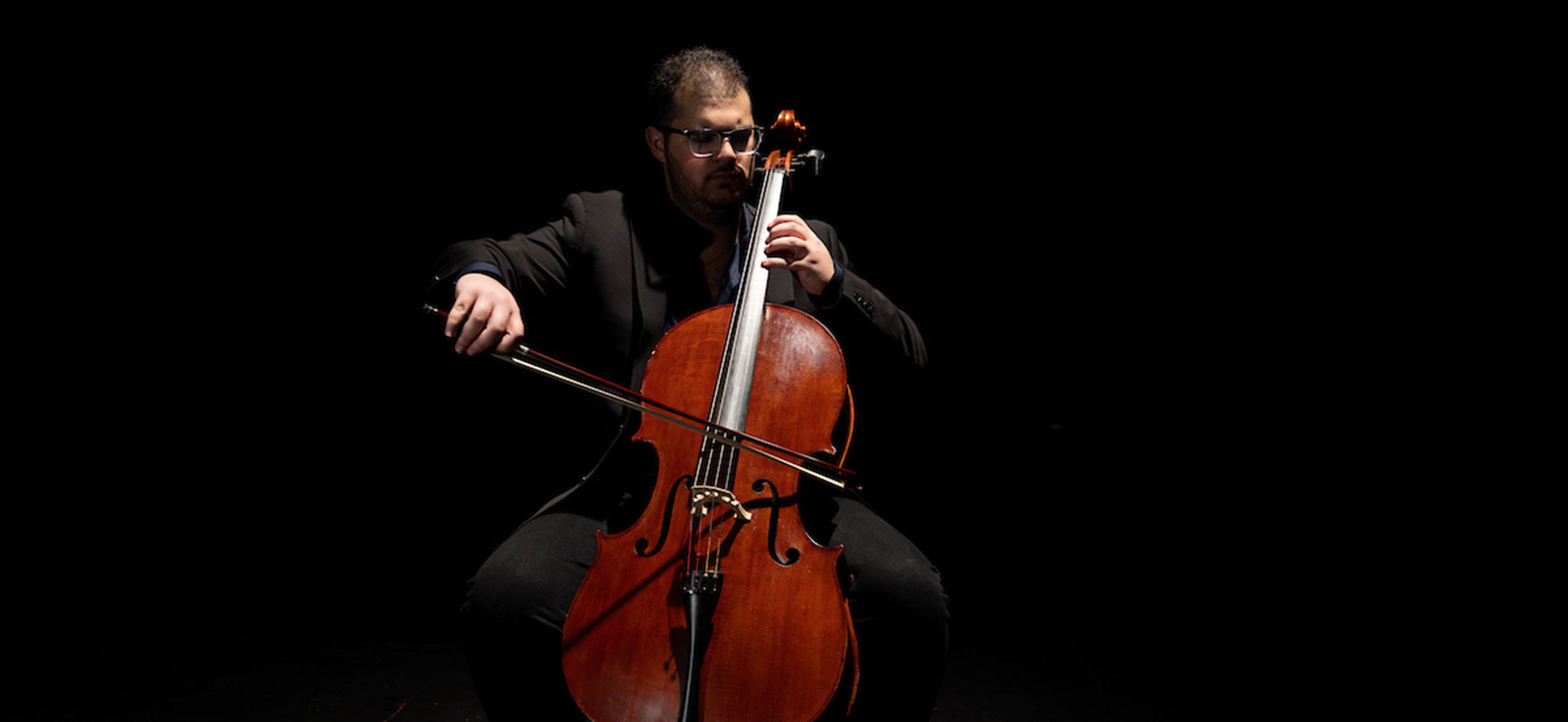 Bryan da Costa plays a cello on a dark stage.