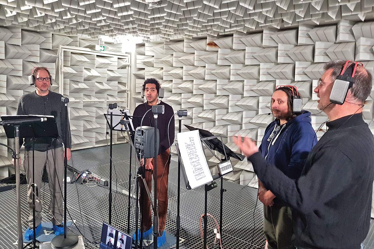 Four people wear headphones and sing in a room with white, geometric walls.