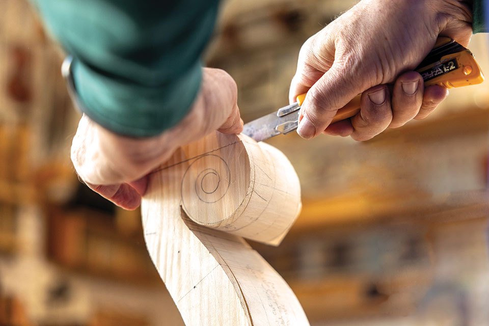 Hands carving a cello's scroll with a knife.