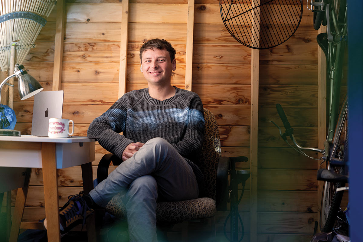 Sam Wachman sits cross legged in his workspace; on the table beside him are a cup of coffee and a laptop.