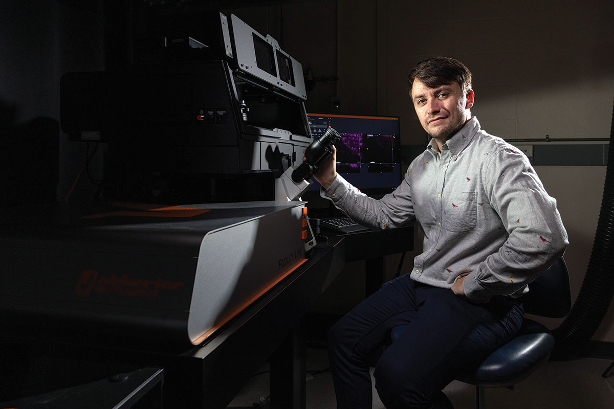 Andy Stone sitting in a dimly-lit lab next to Brandeis’ STED microscope.