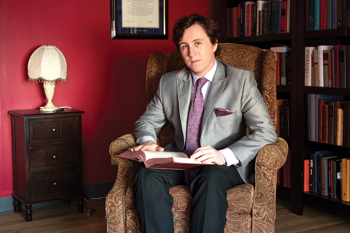 Nathan Robinson wears a suit while sitting in a chair in an office; a wall of books are behind him and a book is open on his lap