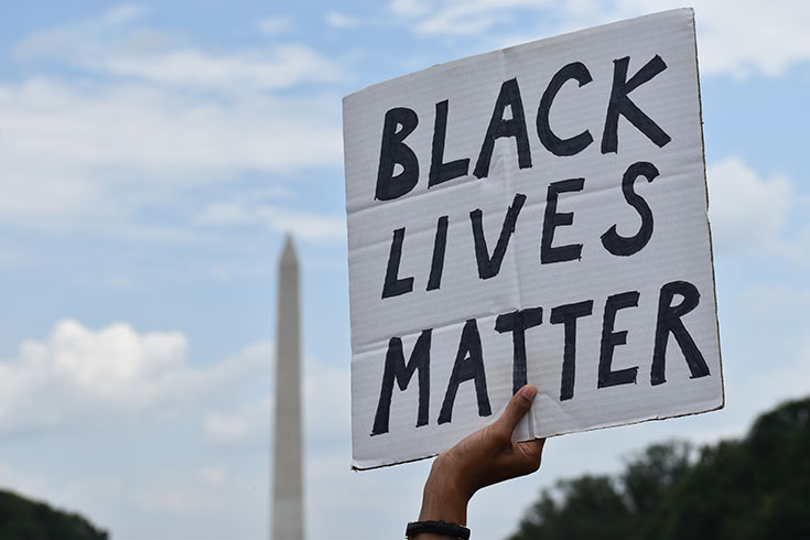 A hand holds a sign that reads "Black Lives Matter" in front of the Washington Monument.