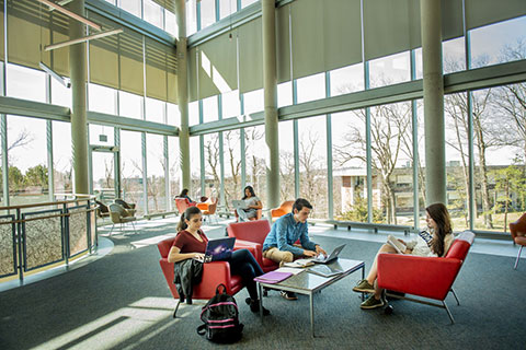 Students sitting in chairs around a small table while reading and working on laptops surrounded by windows in the brightly lit Mandel Center.