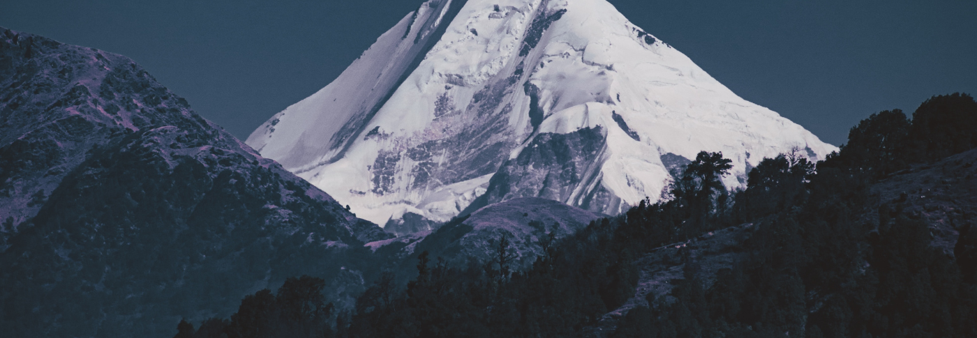 A mountain peak in snow with dark foreground and lower peaks