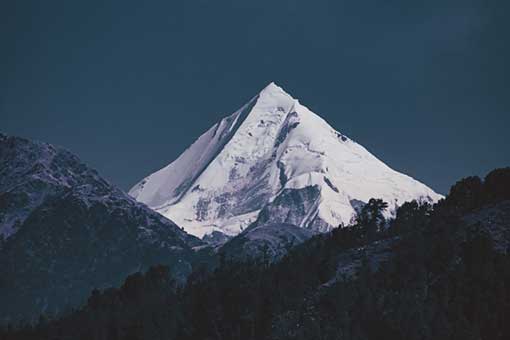 A mountain peak in snow with dark foreground and lower peaks