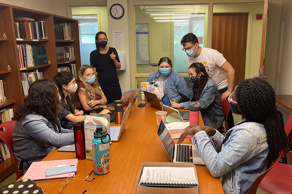Prof. Ziva Hassenfeld stands in a conference room addressing a group of her student researchers sitting around a table. All are wearing masks.