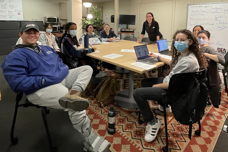 Professor Hassenfeld stands addressing a group of students seated around a large wooden table with many black chairs around it and a patterned rug underneath. In the background, there is a whiteboard, a screen with students on Zoom, a lamp, and a plant. All people are wearing masks. The table is covered in laptops and papers.