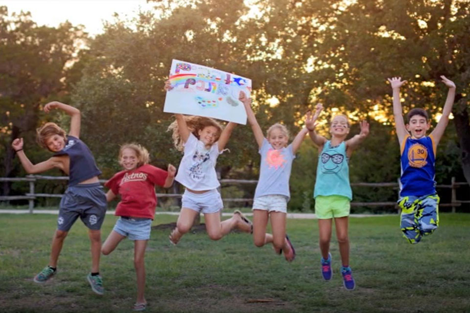 Screenshot from movie trailer for the book "Hebrew Infusion: Learning and Community at American Jewish Summer Camps" with picture of kids jumping high in the air holding a hand made sign with Hebrew writing.