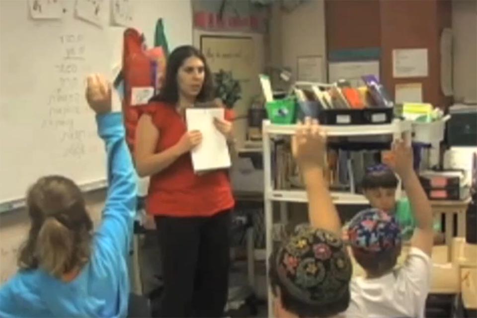 Jocelyn Siegel standing at the front of her classroom posing the opening question.  Students are raising their hands to answer.