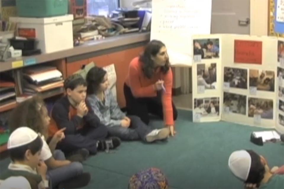 Teacher kneeling on the rug with students sitting in a circle, all looking at a bulleting board with pictures