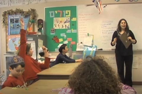 Teacher standing at the board talking to a class of young students.  One girl is raising her hand high.