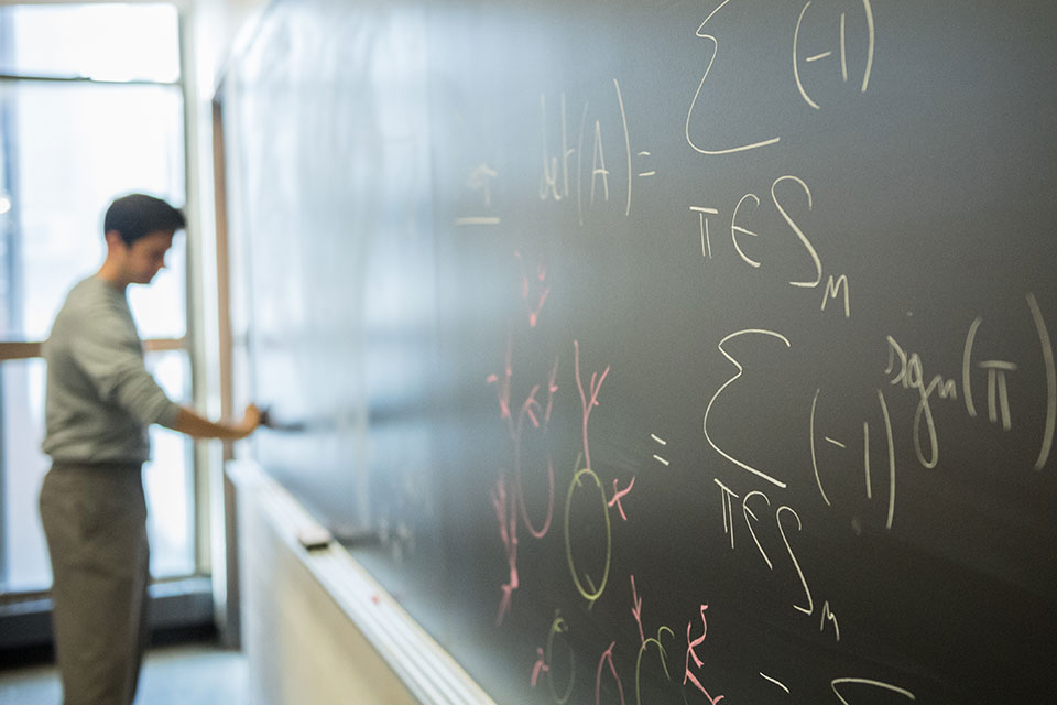 A close up of a chalkboard, with a professor writing on it in the background