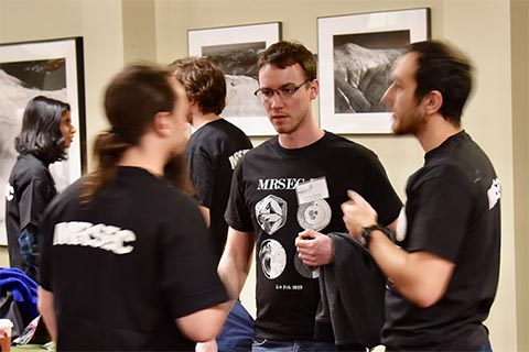 Three students engaged in discussion.  In the background are people looking at posters.