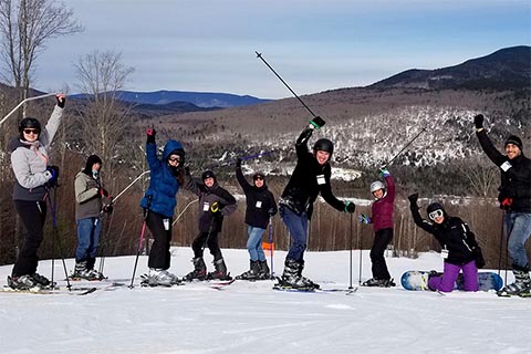 8 skiiers and one snowboarder pose for a picture by a scenic vista. Several hold their ski poles or their fist up in the air with big smiles.