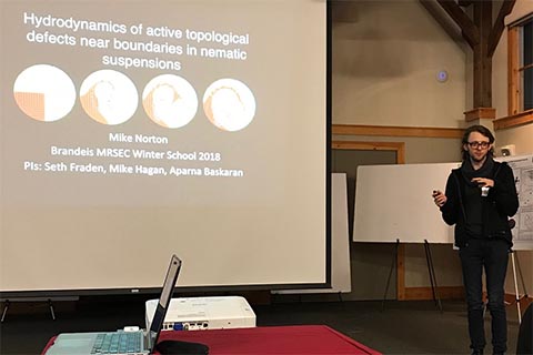 Mike Norton delivering a presentation.  Slide text: Hydrodynamics of active topological defects near boundaries in nematic suspensions.  Mike Norton.  Brandeis MRSEC Winter School 2018.  PIs: Seth Fraden, Mike Hagan, Aparna Baskaran." 