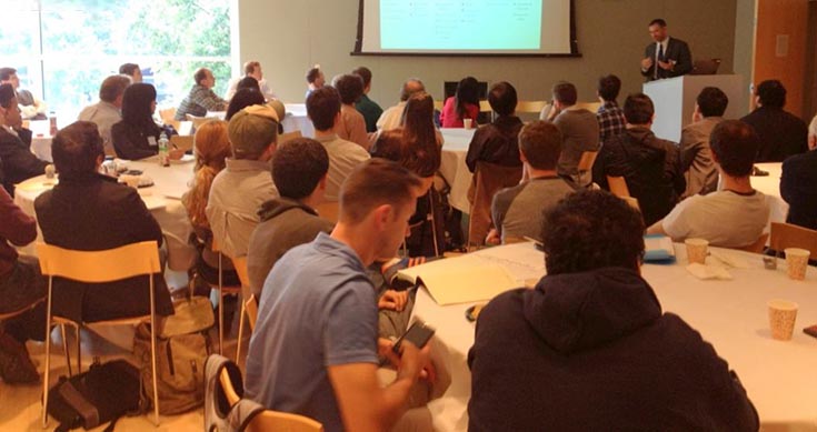 Large room at a symposium with presenter at the podium speaking, and many attendees at tables.