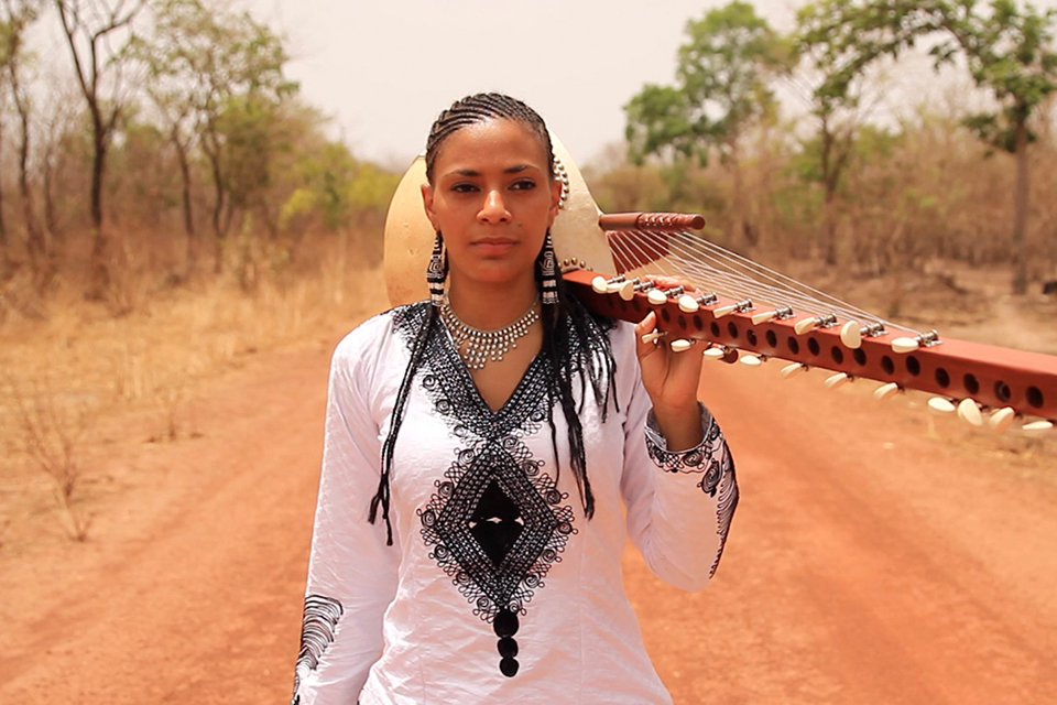 Sona Jobarteh walks down a dusty path holding her kora