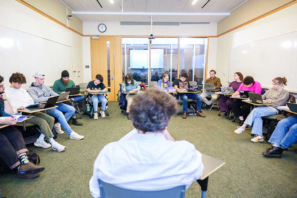  Balding grey headed man sits in front of a classroom.