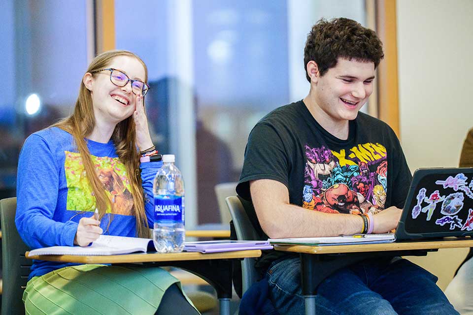 Two students sitting at desks, man and woman, laughing.