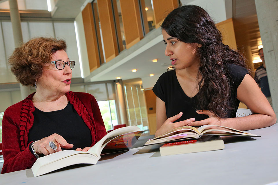 A female professor talks with a student, both with books open in front of them on a table