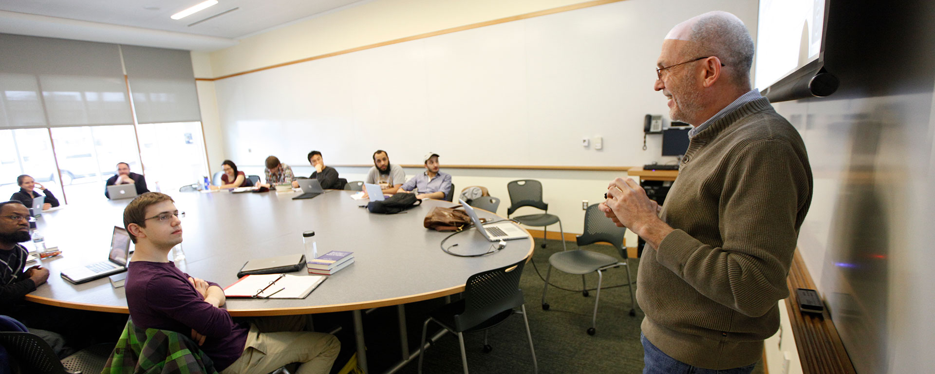 A professor speaks to a group of students sitting around a table in class