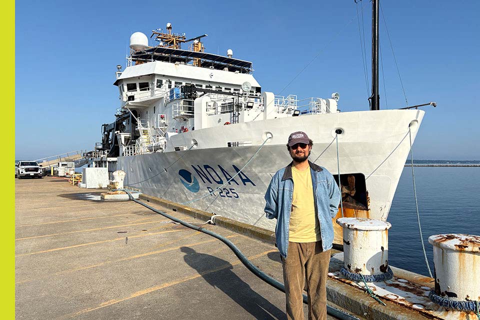 Greg stands on a dock, a NOAA ship is seen behind him