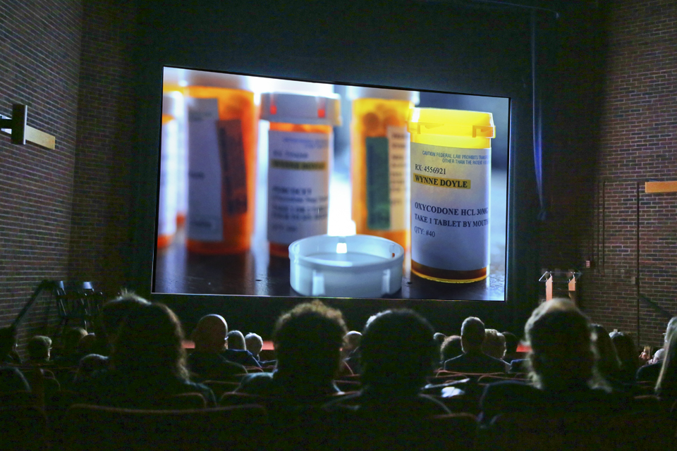 audience in darkened theater look at bottles of prescription painkillers, Ocycodone is the bottle in focus