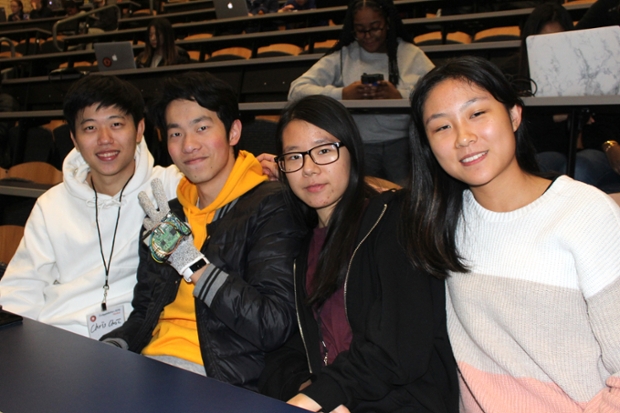Four stidents sit in a lecture hall looking at camera - one wears a glove and gives a peace sign