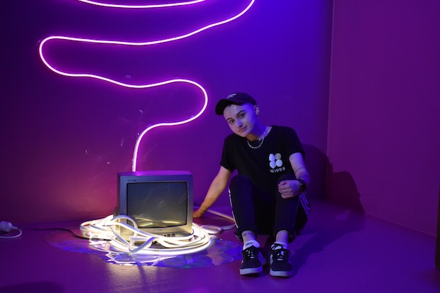 Nico Leger poses next to an art installation featuring a small old TV and fluorescent lights. 