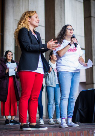A woman in red pants and blue blazre applauds next to Linzy Rosen, speaking with microphone in white t-shirt and jeans