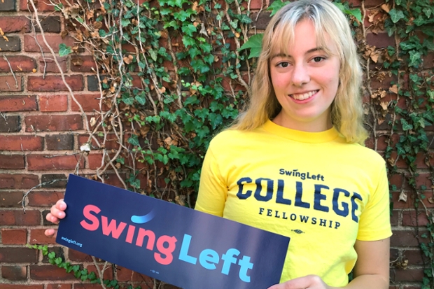 Holly Newman '22, in a yellow t-shirt with a blue "swing left" bumpersticker, standing in front of an ivy-covered brick wall