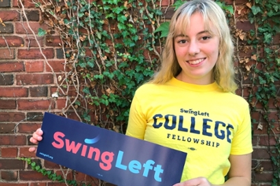 Holly Newman '22, in a yellow t-shirt with a blue "swing left" bumpersticker, standing in front of an ivy-covered brick wall