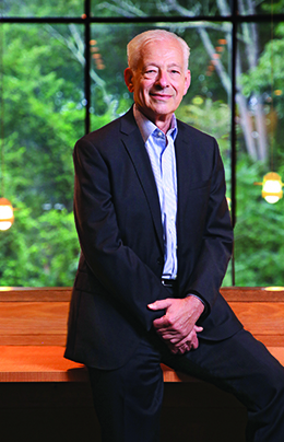 Professor Peter Petri in suit with no tie leaning against a wooden counter with green trees visible through a window behind him