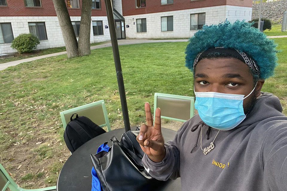 Murad Anderson '23, with teal hair, face mask and sweatshirt, gives the peace sign at a table in the Ziv quad