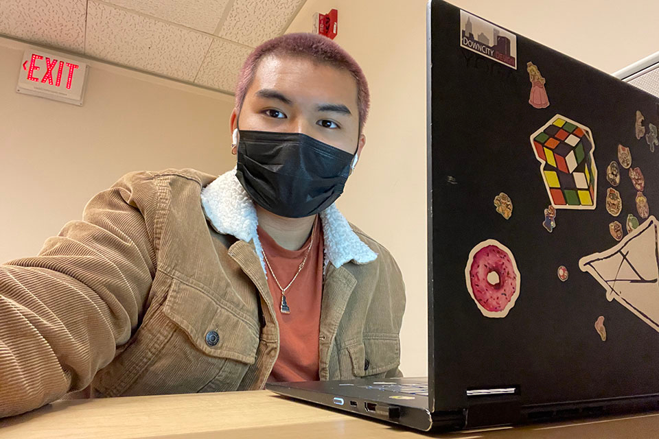 Nick Ong, with a pink buzz cut, black face mask and suede sheerling jacet in front of his laptop in the basment of the Library