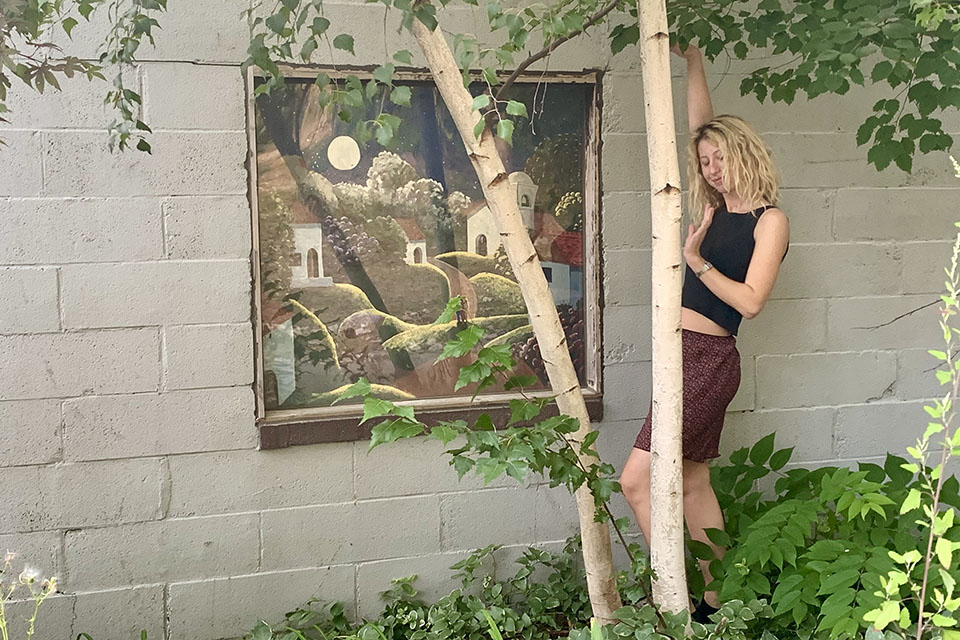 Solvai Lewenberg, '23, stands next to a birch tree in front of a mural on the Women's and Gender Studies building