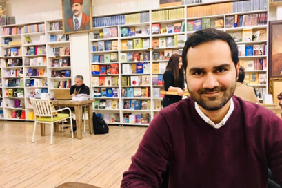 Houman Oliaei sits at a table in the right of the frame in front of open shelves of books.