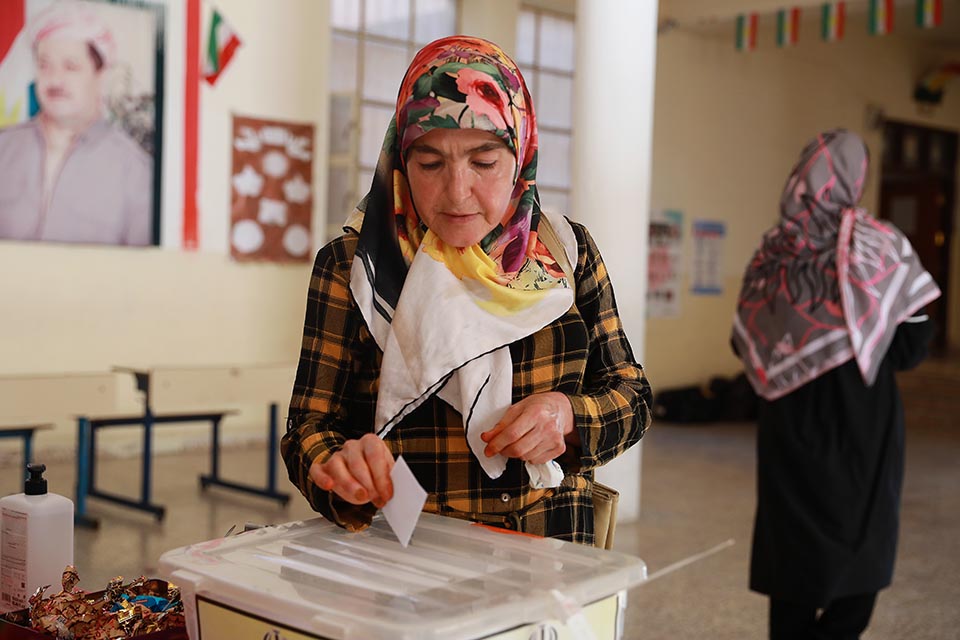 A woman in a floral hijab and plaid jacket places a ballot into a box in Iraq