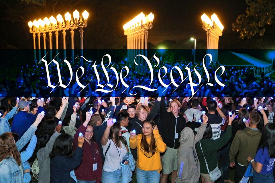 An image of students at the Light of Reason at night, with a blue banner superimposed over it with the words "We the People" in calligraphy