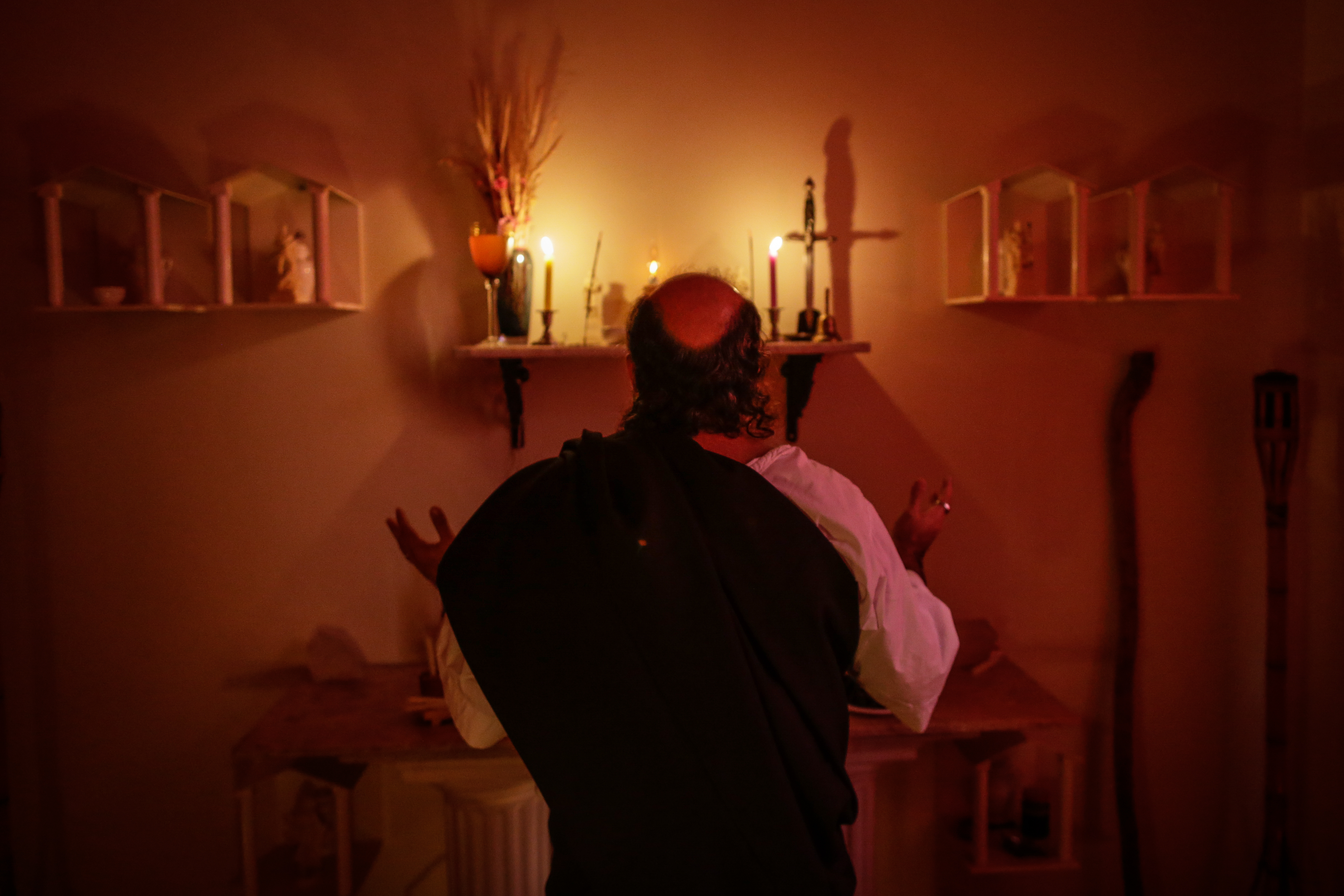 A Wiccan priest stands in front of an alter in a candlelit room. His back is to the camera.