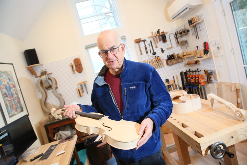 Hairy Mairson stands in his workshop surrounded by tools, holding a partially-made violin 