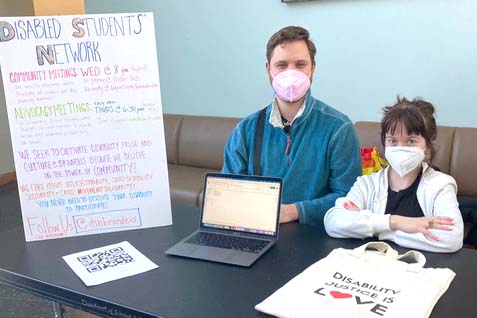 Luca Swinford ‘22 sits in the Shapiro Students Center next to Zoe Pringle '22, and a sign for their club, the Disabled Students' Network.