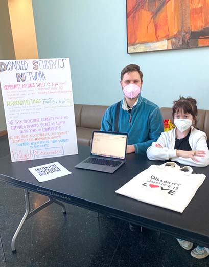 Luca Swinford ‘22 sits in the Shapiro Students Center next to Zoe Pringle '22, and a sign for their club, the Disabled Students' Network.