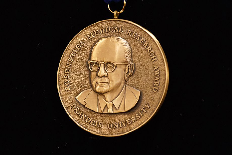Round, gold-colored medallion with the words " Rosenstiel Medical Research Award Brandeis University" circling the head of a man wearing glasses and a tie. Medallion is against a black background.